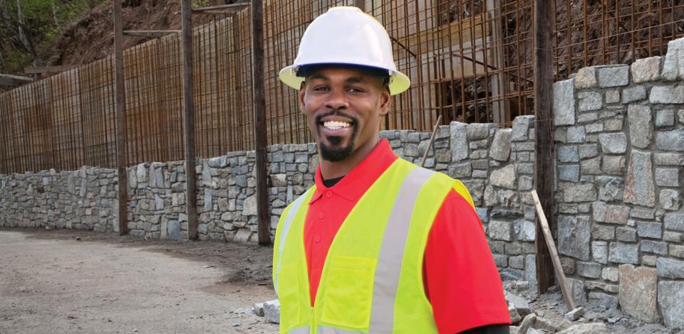 A man wearing a safety vest and hard hat standing in front of a stone wall in construction. 