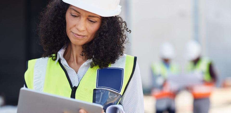 Young female construction engineer wearing a safety vest standing outdoors on a worksite using a digital tablet with workers in the background